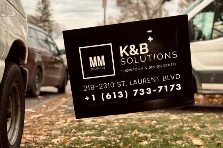 Dark MM Brothers K&B Solutions lawn sign with white lettering beside parked vehicles.
